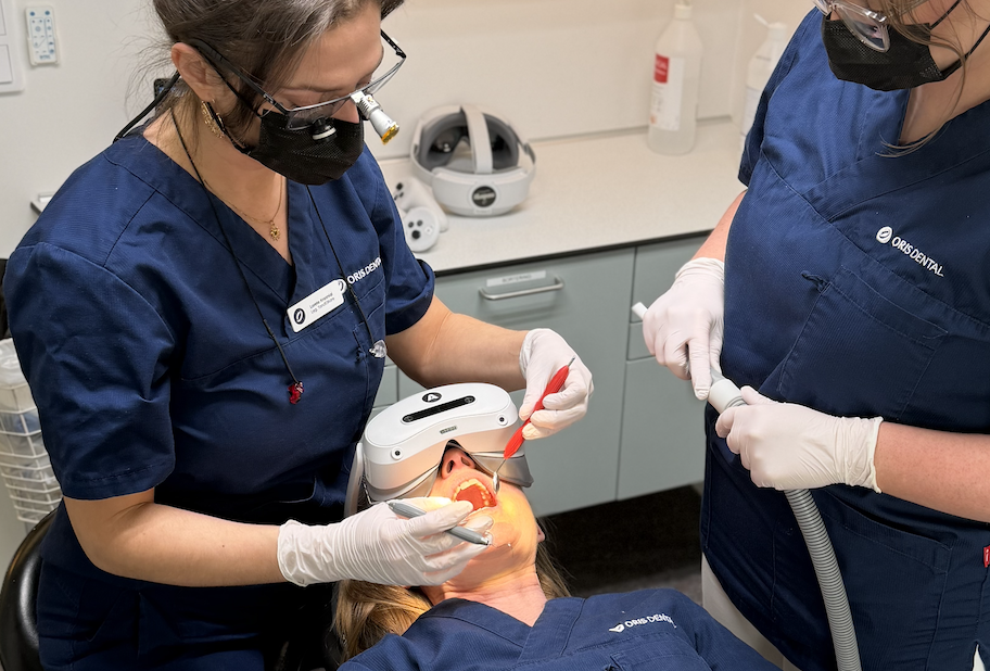 Image of two dentists at oris dental working in another persons mouth. The patient is wearing Mindergy VR headset in the dental chair.
